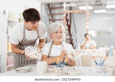 Positive young guy assisting enthusiastic senior woman painting ceramic mug in pottery class, showing plate with paint samples. Intergenerational communication and creative hobby concept - Powered by Shutterstock