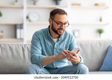 Positive young guy in eyeglasses sitting on sofa with mobile phone and texting to his friend, indoors. Millennial Caucasian man writing message, having video call or broswing web at home