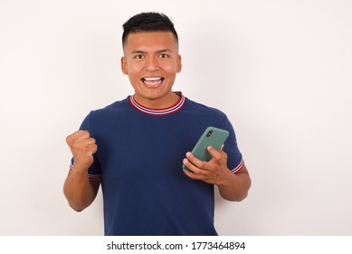 Positive Young handsome hispanic man wearing casual t-shirt standing over white isolated background holds modern cell phone connected to headphones, clenches fist from good emotions, exclaims with joy - Shutterstock ID 1773464894