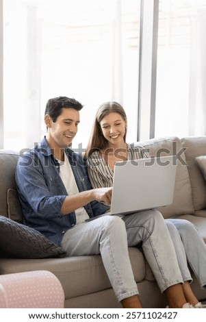 Positive young Hispanic couple using online application on laptop for media entertainment at home, enjoying modern Internet technology, shopping on ecommerce platform together. Vertical shot