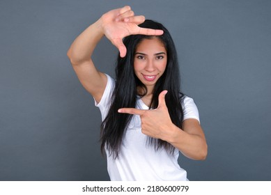 Positive Young hispanic  woman wearing white T-shirt over grey background with cheerful expression, has good mood, gestures finger frame actively at camera. - Powered by Shutterstock