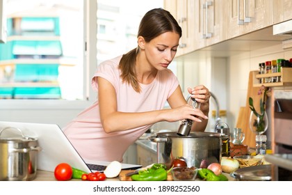 Positive young housewife cooking vegetable soup and using laptop at kitchen