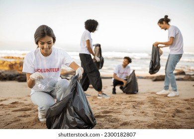 Positive young international people volunteers in gloves and european lady with garbage bags clean up trash on beach, outdoor. Eco and environment conservation protecting planet, ocean pollution - Powered by Shutterstock