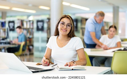 Positive young lady in casual clothes and glasses working remotely using a computer in a quiet library - Shutterstock ID 2245801121
