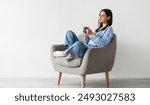 Positive young lady in casual wear sitting in armchair with crossed legs, warming hands on cup of hot coffee, enjoying warm drink against white studio wall, copy space