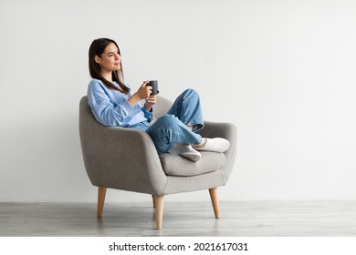 Positive young lady in casual wear sitting in armchair with crossed legs, warming hands on cup of hot coffee, enjoying warm drink against white studio wall, copy space - Shutterstock ID 2021617031