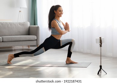 Positive Young Lady In Sportswear Standing In Warrior Pose, Watching Online Yoga Class, Yoga Coach Shooting Video For Her Students Or Followers, Using Smartpohne On Tripod, Home Interior