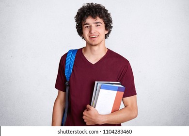 Positive young male teenager with pleasant charming smile, holds books, carries bag, prepares for classes at college, likes studying and find out something new, isolated on white background.