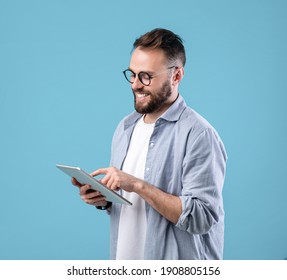 Positive Young Man In Glasses Using Digital Tablet For Online Work Or Studies On Blue Studio Background. Happy Teacher Giving Webinar. Blogger Writing New Post For Social Media