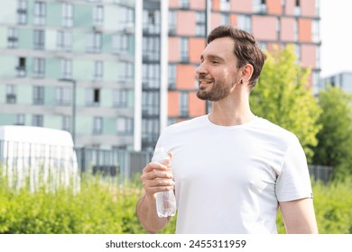 A positive young man has jogging in a modern residential area with colorful buildings and parkland on a summer morning. A smiling sportsman stands sideways, takes a break, looks forward and drinks - Powered by Shutterstock