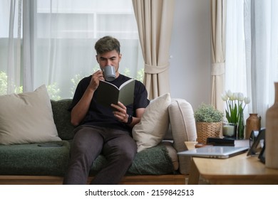 Positive young man reading book and drinking coffee while sitting on sofa at home. - Powered by Shutterstock