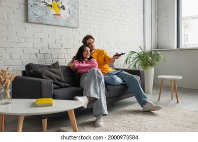 positive young man sitting on couch, holding remote controller and hugging girlfriend in modern living room