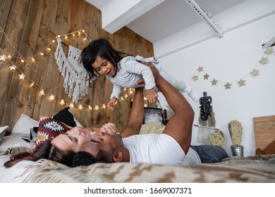 Positive Young Mixed Race Family Cute Young Mother African American Dad And Charming Smiling Daughter Have Fun On The Bed On Friday Evening. Family Time Concept