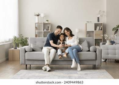 Positive young mom, dad and sweet girl kid taking family selfie on cozy comfortable couch, having fun with digital gadget in cozy stylish home interior, using smartphone for selfie together - Powered by Shutterstock