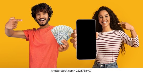 Positive young multiethnic couple recommending nice gambling mobile app, pointing at cell phone with black blank screen and cash dollars on yellow studio background, mockup, collage, panorama