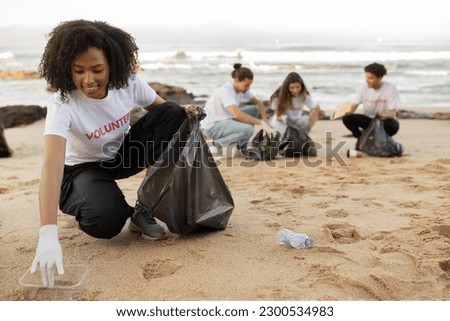 Positive young multiracial people volunteers in rubber gloves and african american lady with trash bag clean up garbage on sea beach, outdoor. Eco, environment conservation, protecting planet