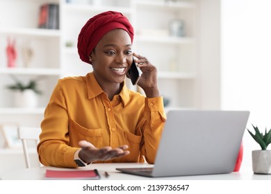 Positive Young Muslim Black Lady With Red Turban On Her Head And Casual Outfit Working On Laptop At Office, Looking At Computer Screen And Talking On Cell Phone With Business Partner, Copy Space