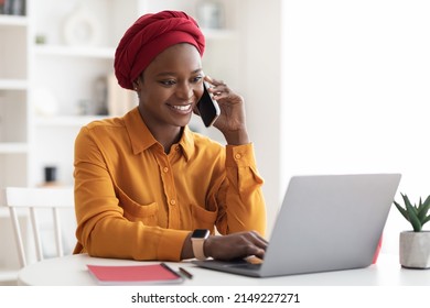 Positive Young Muslim Black Woman With Red Turban On Her Head And Casual Outfit Working On Laptop At Office, Looking At Computer Screen And Talking On Smartphone With Business Partner, Copy Space