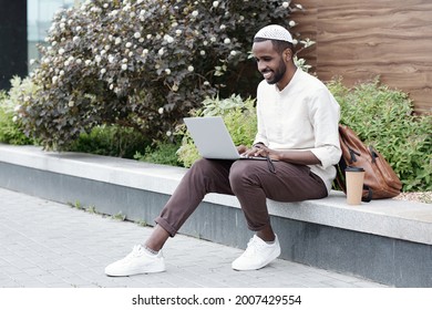 Positive Young Muslim Marketer In Kufi Cap Sitting On Curb And Using Laptop While Working Online