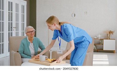 Positive Young Nurse Carries Tray With Breakfast To Senior Woman Patient Sitting In Light Living Room