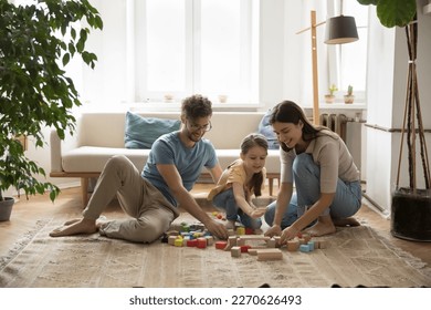 Positive young parents and cute kid playing with wooden colorful toy blocks on warm carpeted floor together, relaxing in living room. Mom and dad playing with girl at home - Powered by Shutterstock