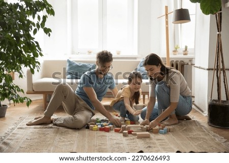 Positive young parents and cute kid playing with wooden colorful toy blocks on warm carpeted floor together, relaxing in living room. Mom and dad playing with girl at home