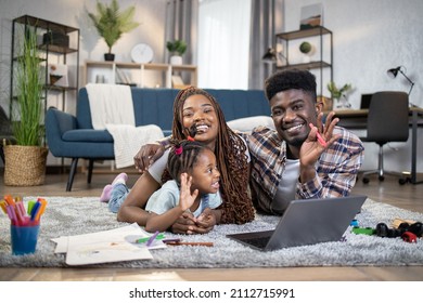 Positive Young Parents Embracing Pretty Daughter While Lying On Floor With Modern Laptop, Toys And Pencils. Happy African Family Waving Hands And Smiling On Camera.