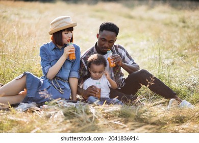 Positive Young Parents And Their Cute Son Sitting Together On Grass And Drinking Fresh Juice From Straw. Adorable Multi Ethnic Family Enjoying Picnic Time During Summer Days.