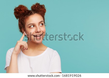 Positive young pretty curly brunette woman with bun hairstyle keeping raised forefinger on her earphone and looking aside with pleasant smile, standing over blue background
