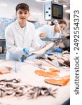 Positive young salesman demonstrating seabass behind counter in fish store with large assortment