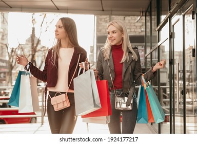 Positive Young Stylish Female Friends In Trendy Outfits Smiling While Walking On City Street With Shopping Bags