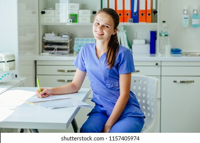 Positive Young Woman Assistant Sitting On Workplace Of The Medicall Office In Gynecological Clinic. Female Medical Doctor Writing Something Sitting At Her Office. Healthcare And Medical Concept.