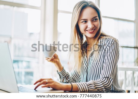 Positive young woman copywriter with cup of tasty coffee in hand looking away sitting at modern laptop computer and working freelance in coffee shop with copy space area for advertising message