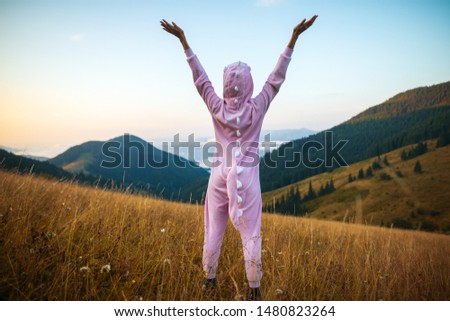 Positive young woman hiker in pink Dragon Pajamas open arms at mountain peak. Slim lady in pink night-suit. Funny travel, Animal Cosplay Costume. Summer adventure journey in mountain nature outdoors