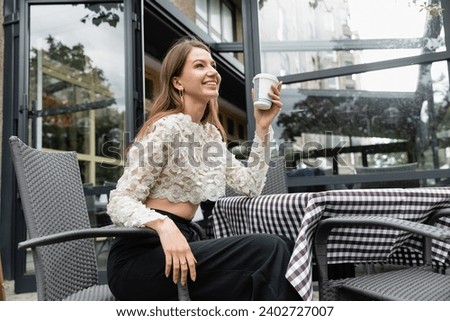 positive young woman holding coffee to go while sitting at table on terrace near cafe in Berlin