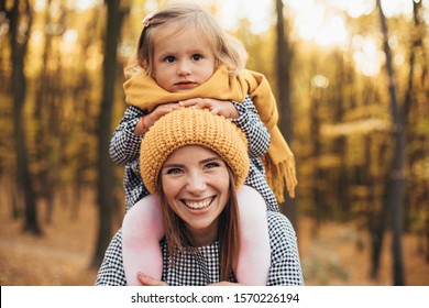 Positive Young Woman Look On Camera And Smile. Her Daughter Sit On Her Neck. Serious Small Girl Look Straight Too. Woman Wear Orange Hat While Girl Has Scarf. Autumn Weather. Stand In Forest Or Park