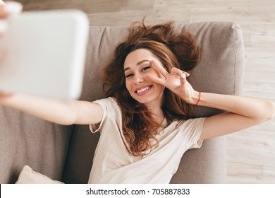 Positive young woman showing peace gesture and smiling while making selfie at home - Shutterstock ID 705887833