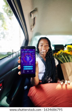 Positive young woman sitting on taxi backseat and showing smartphone with smart charger application