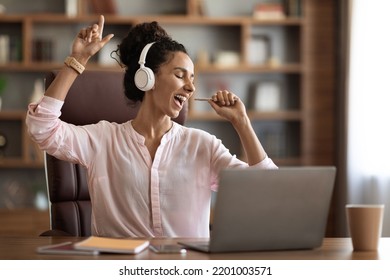 Positive young woman sitting at workdesk in front of computer, using wireless headset and pen as microphone, freelancer listening to music and singing while having break, copy space. Stress relief - Powered by Shutterstock