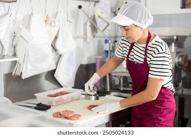 Positive young woman in striped shirt, hairnet and apron, pressing fresh raw ground beef hamburger patties for sale in local butcher shop .. - Powered by Shutterstock