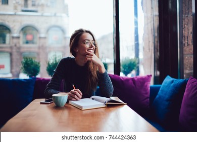 Positive young woman in stylish eyeglasses dreaming while sitting at table with opened book in stylish coworking space.Smiling beautiful female in spectacles looking away during reading bestseller - Powered by Shutterstock