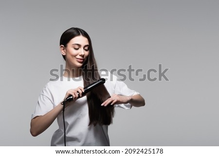positive young woman using hair straightener isolated on grey