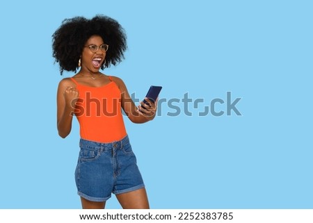 positively surprised young woman celebrates, holds a smart phone in one hand, black power afro style hair on blue background