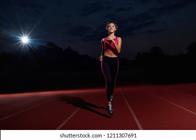 Positivity And Happy Young Woman In Sport Wear Running Forward On Track, Looking And Smiling At Camera. Athletic Girl With Curly Hair Training, Preparing Alone To Competition On Stadium At Night Time.