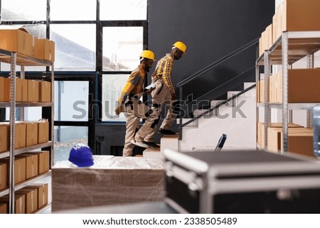 Post office storage workers going upstairs to warehouse second floor, carrying barcode scanner and tablet. Smiling african american industrial storehouse colleagues chatting and working together