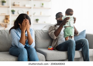 Post Partum Depression. Exhausted Black Mom Covering Face Crying Sitting On Couch At Home, Feeling Headache And Stress, Her Husband Playing With Small Infant Kid. Challenges Of Parenthood. Baby Blues