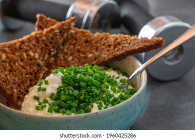 Post Or Pre Workout Meal With Low Fat Cottage Cheese And Wholegrain Bread Served With Chives In A Bowl And Dumbbells In The Background