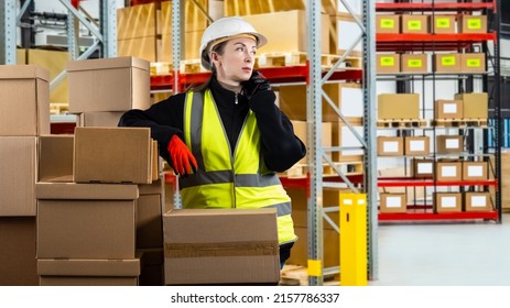 Postal warehouse worker. Woman with phone. Woman works in warehouse for postal company. Cardboard parcels in front of girl with phone. Postal worker is talking on phone. Female in yellow vest