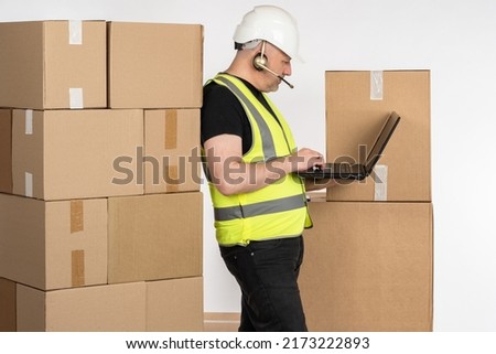 Postal worker with parcels. Postal worker in headphones with microphone. Man in yellow vest and hardhat. Postal worker is holding clipboard. Corton boxes for delivery to client. Delivery business