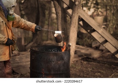 Post-apocalypse. A Man In A Gas Mask Holds A Tin Can Over A Fire Lit In A Rusty Iron Barrel. Selective Focus